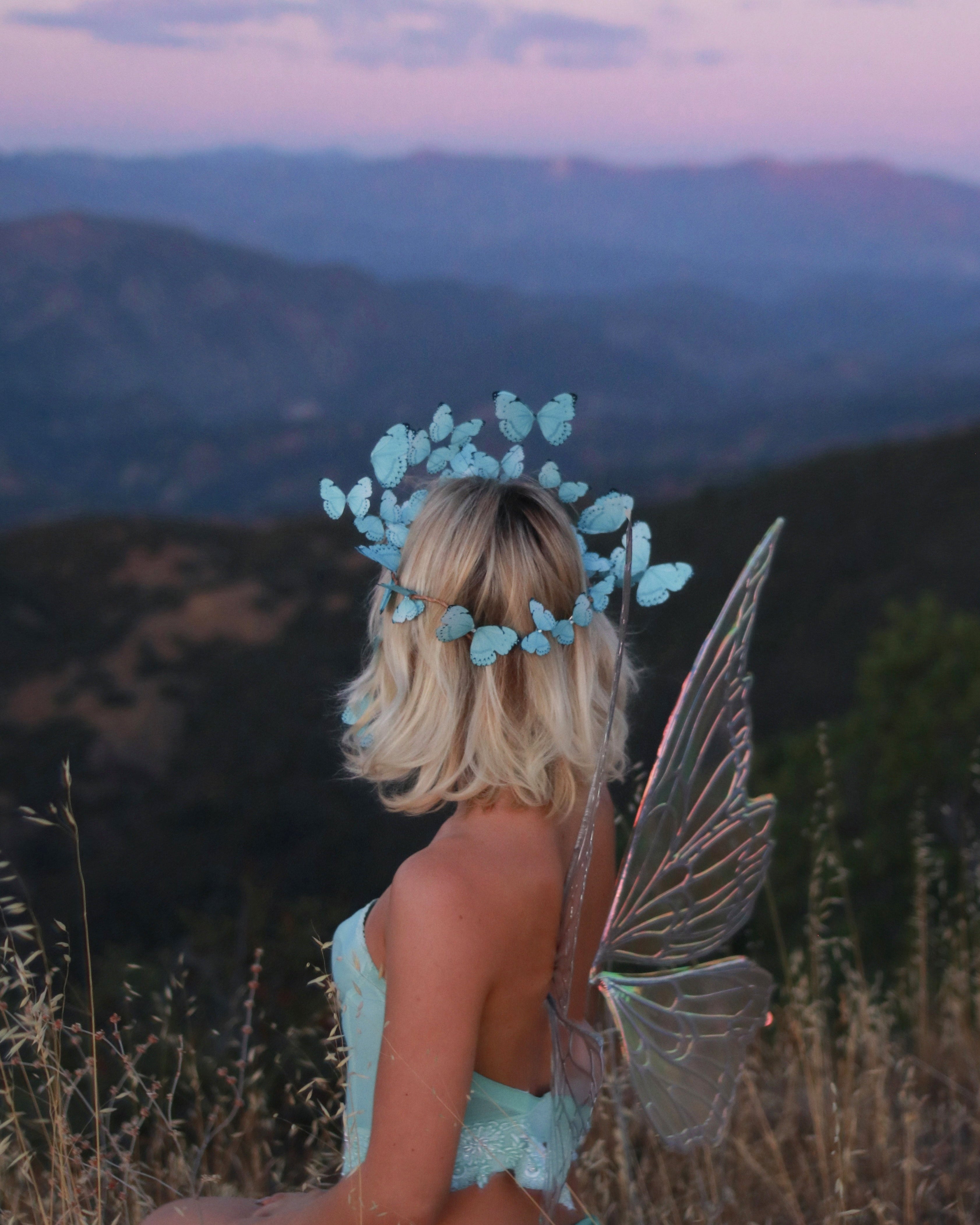 Person in fairy costume with blue flower headpiece and wings standing on a mountain at sunset.