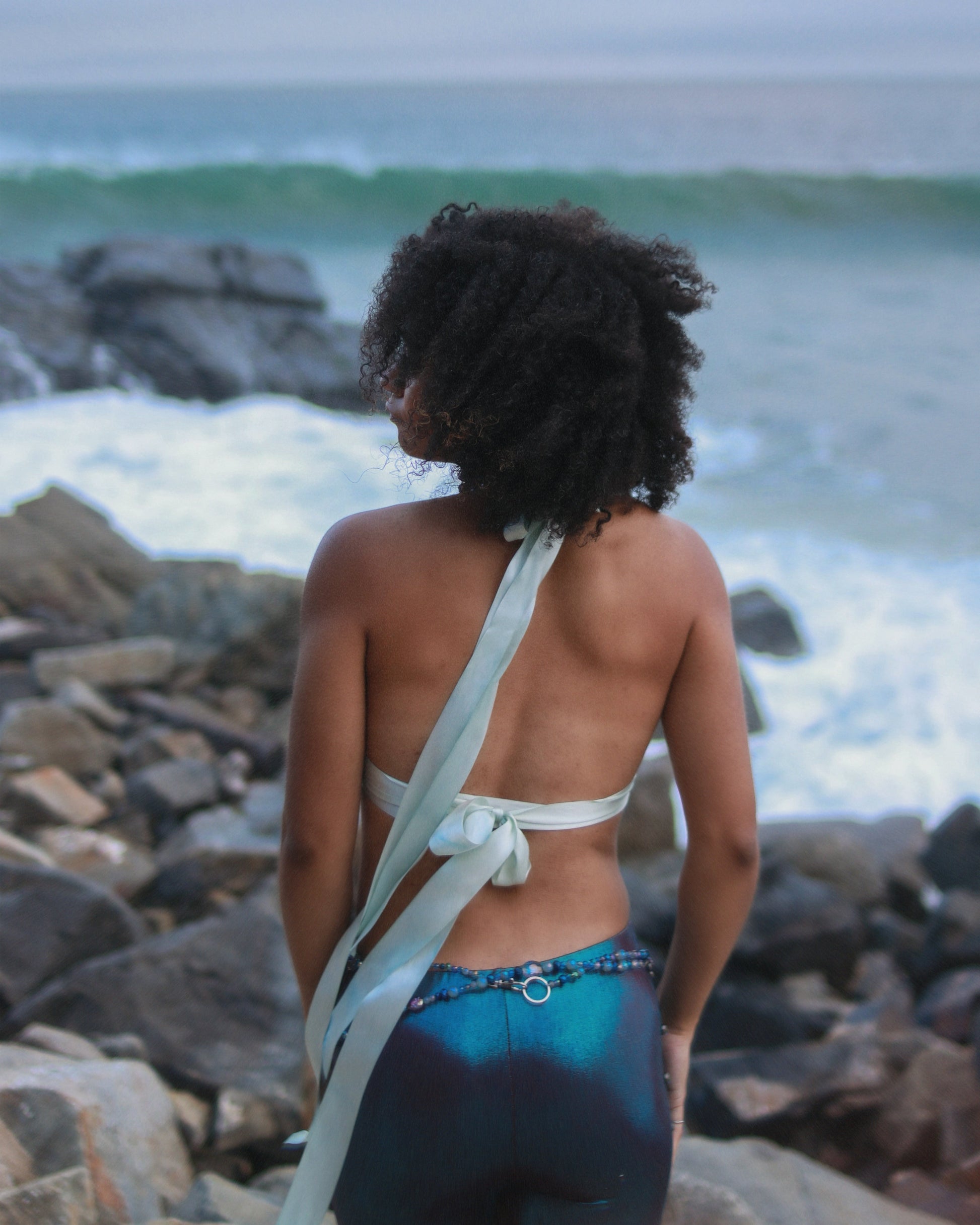 Person with curly hair standing on a rocky beach with ocean waves in the background