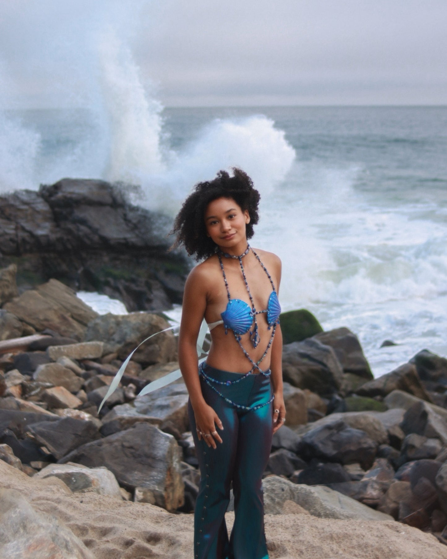 Person wearing a blue bikini top and green pants standing on rocky coastal terrain with ocean waves in the background.