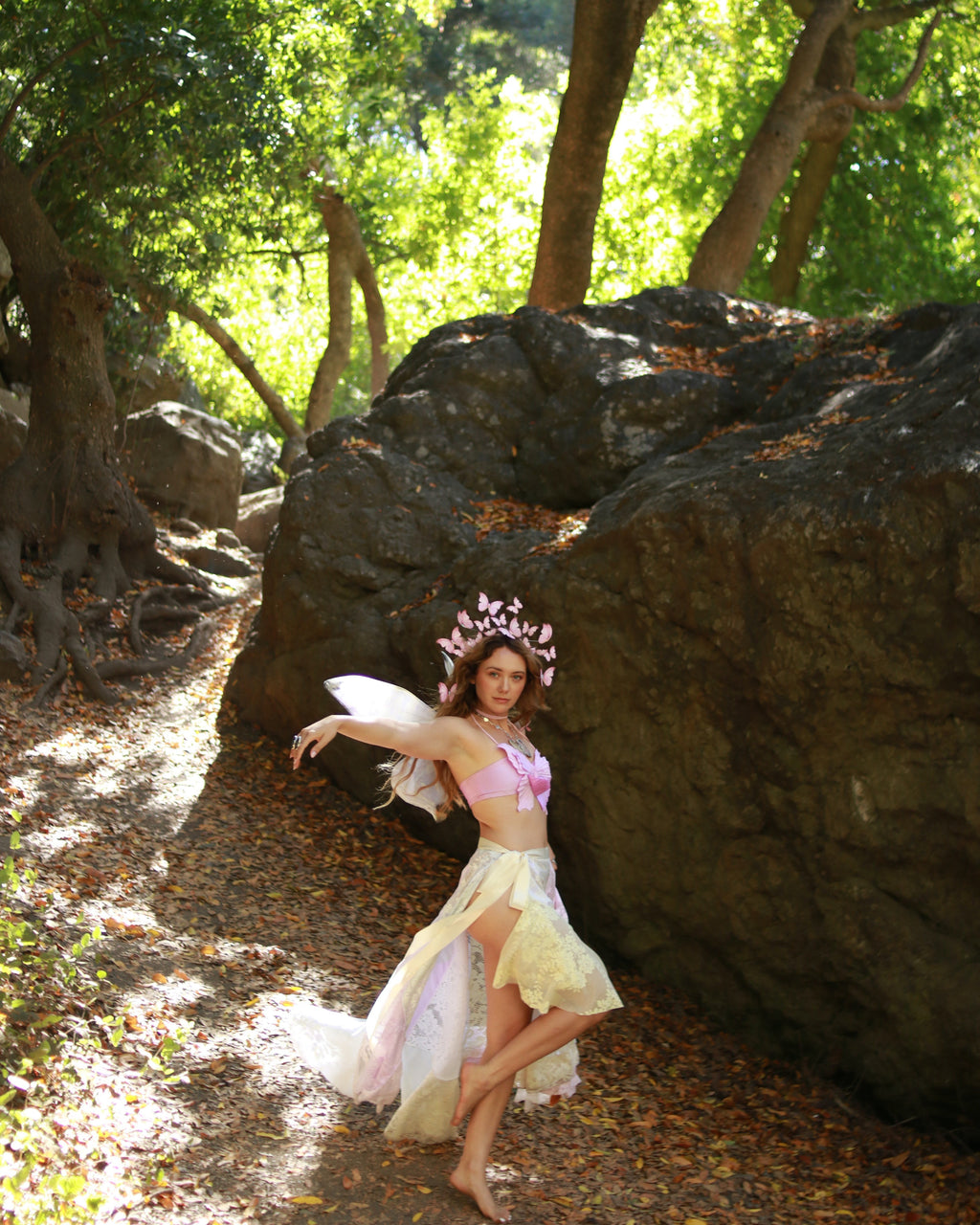 Woman in a fairy costume standing in a forest with large rocks and trees.